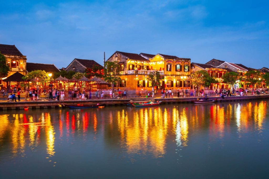 Hoi An Ancient Town waterfront lit up at night, with glowing yellow merchant houses, lanterns, and their reflections shimmering on the Thu Bon River.