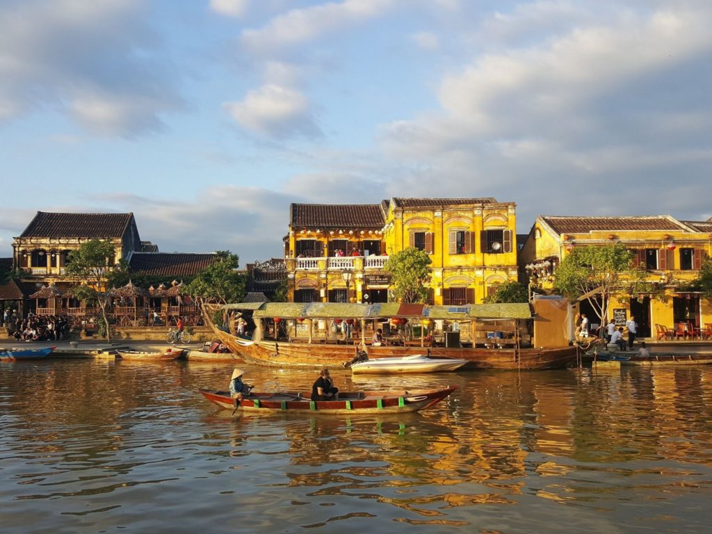 Wooden boats on the Thu Bon River in Hoi An Ancient Town, Vietnam, with iconic yellow merchant houses lining the waterfront at golden hour.