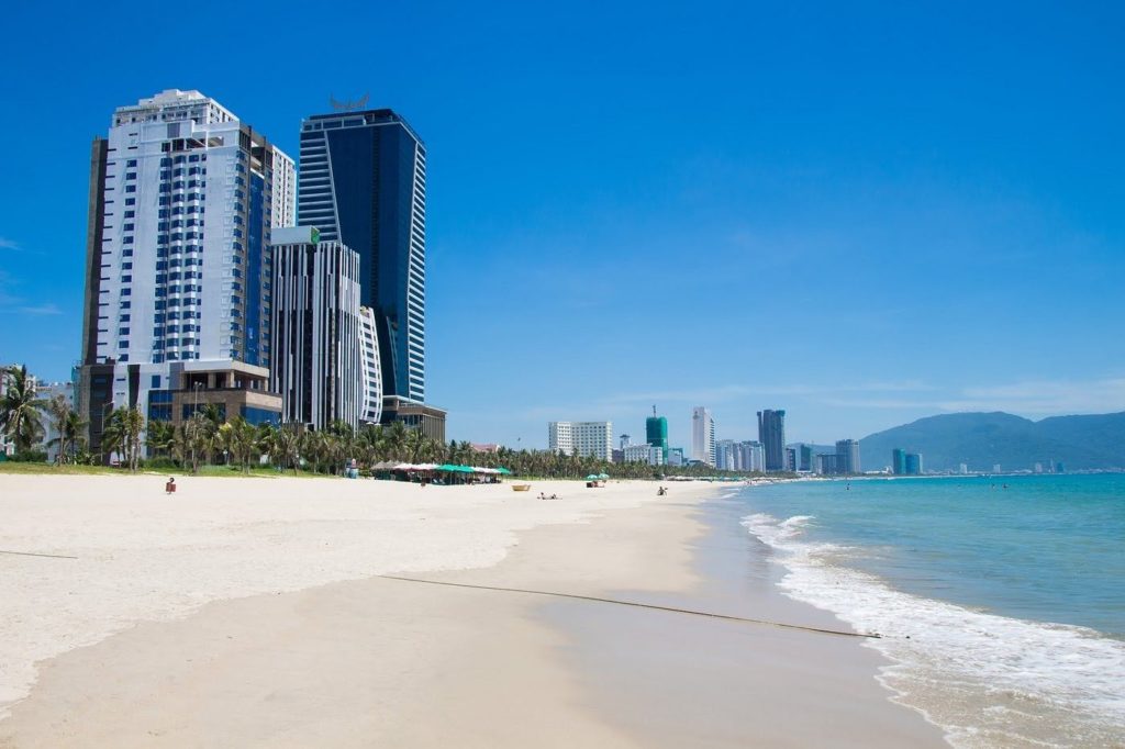 Wide white sand beach at My Khe, Da Nang, with turquoise water, palm trees, and the city skyline stretching along the coast under a clear blue sky.
