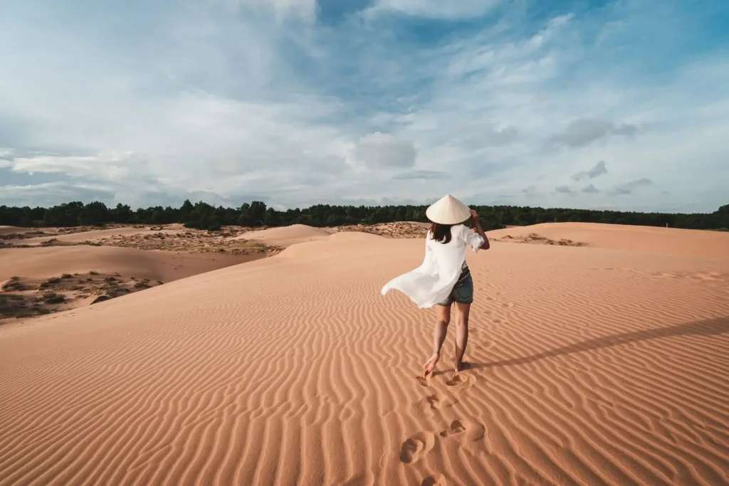 A woman wearing a traditional Vietnamese conical hat and flowing white top walks barefoot across rippled red sand dunes in Mui Ne, Vietnam